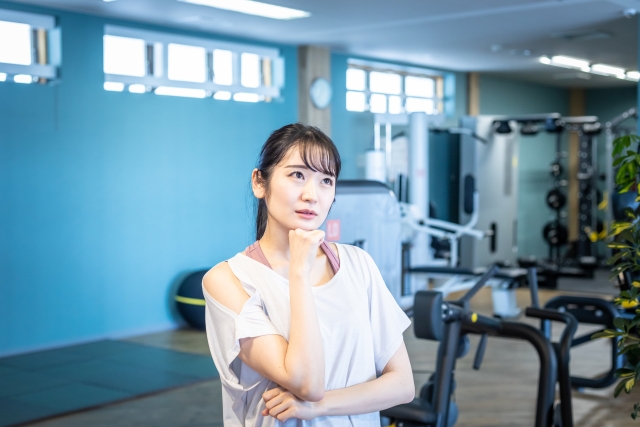 たくさんの筋トレ用マシンが並ぶトレーニングジム（フィットネスジム）で悩む女性